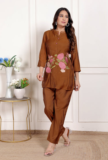 Woman wearing a brown outfit with floral embroidery in a room with a white wall and small table with plants.