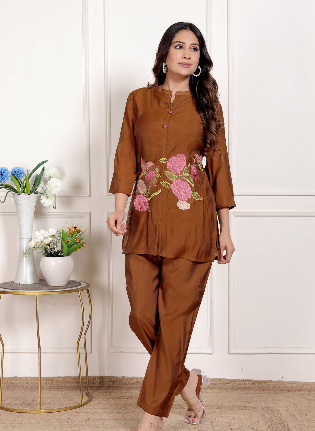 Woman wearing a brown outfit with floral embroidery in a room with a white wall and small table with plants.