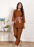 Woman wearing a brown outfit with floral embroidery in a room with a white wall and small table with plants.