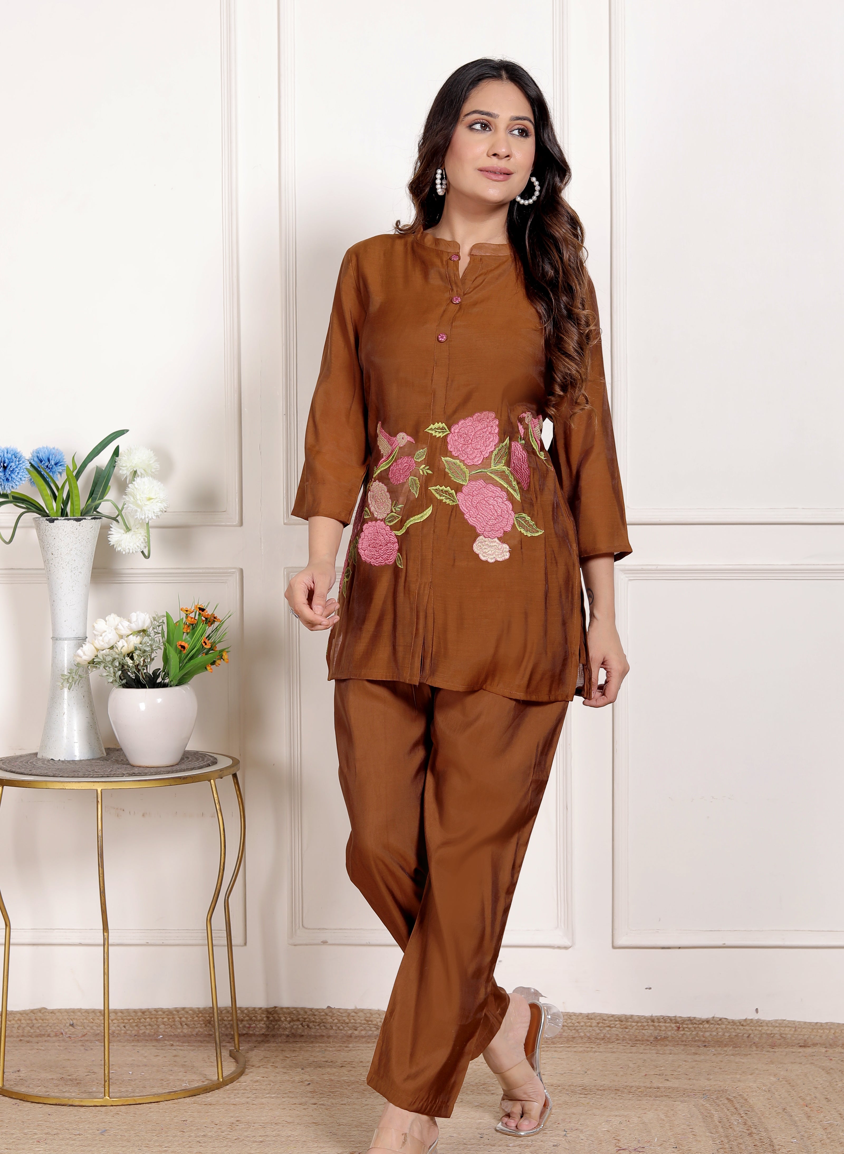 Woman wearing a brown outfit with floral embroidery in a room with a white wall and small table with plants.